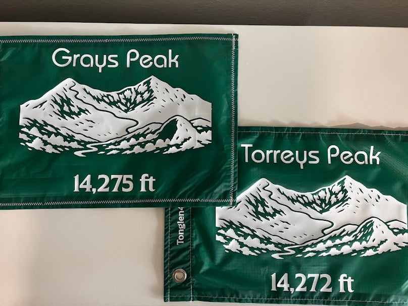 Two green mountain peak flags with white text and illustrations on a neutral background